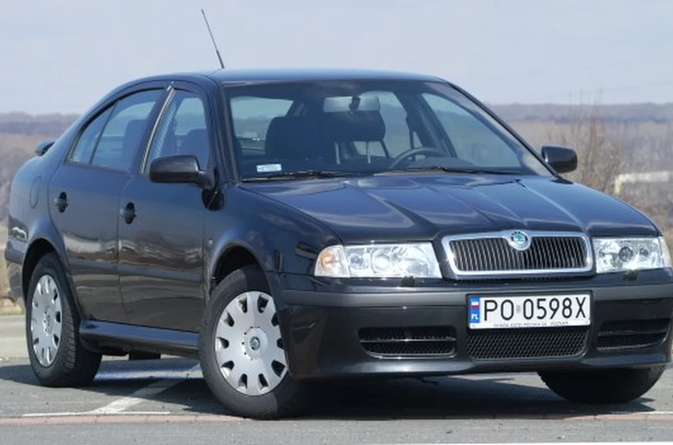 Czarny sedan Škoda Octavia z jasnymi kołpakami, zaparkowany na asfaltowej nawierzchni, posiada widoczną przednią tablicę rejestracyjną o numerze PO 0598X.
