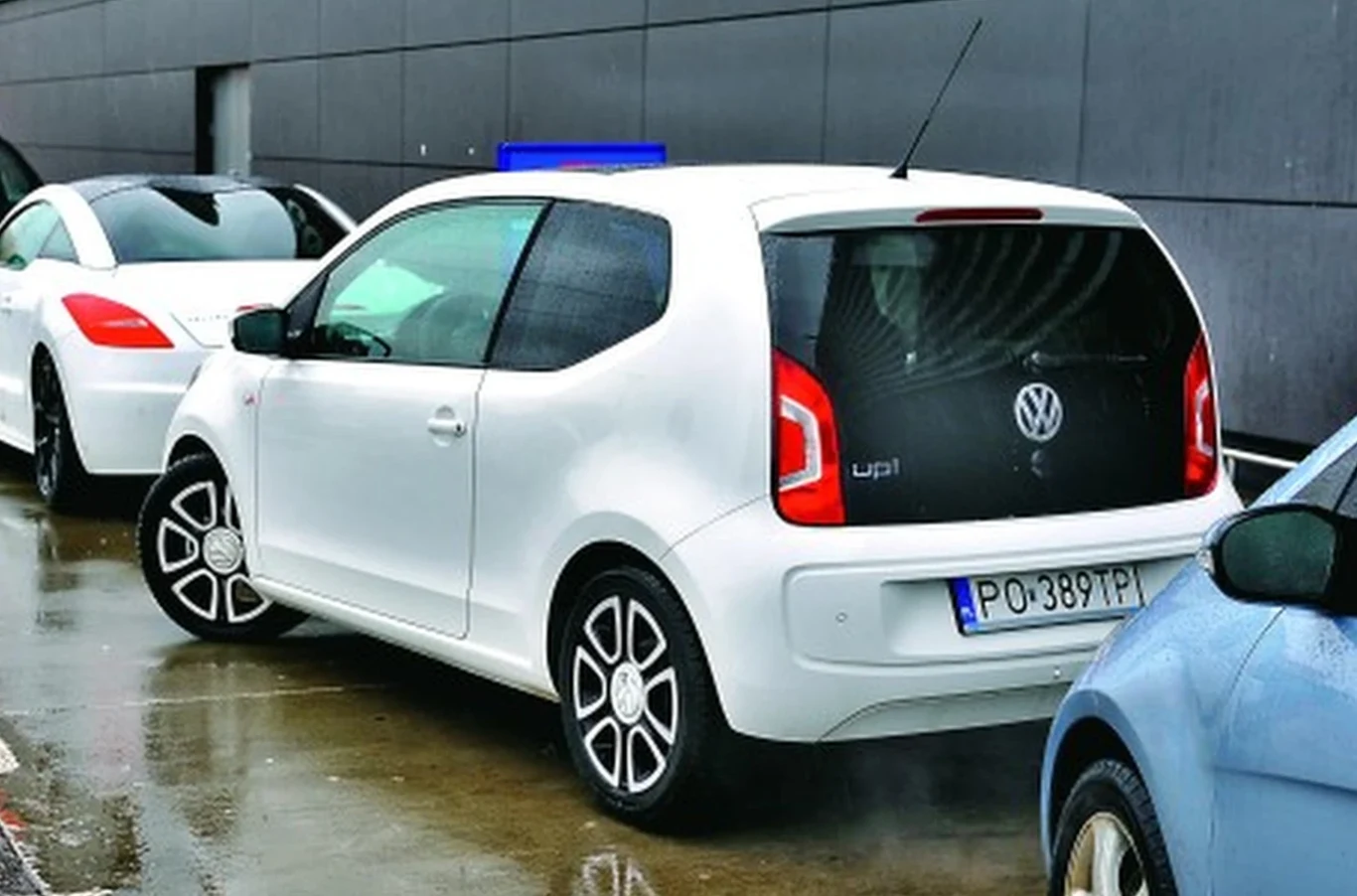 Biały Volkswagen Up! zaparkowany tyłem na mokrym asfalcie, otoczony przez inne samochody w bliskiej odległości przy ciemnej ścianie budynku.