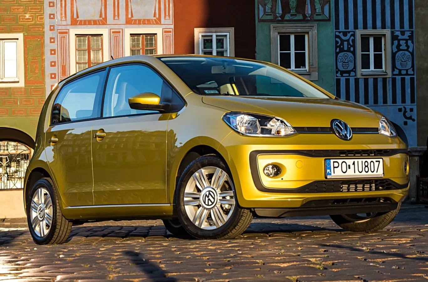 Używane samochody, które mało palą i są tanie w eksploatacji. Żółty Volkswagen Up! zaparkowany na brukowanej ulicy, w tle kolorowe zabytkowe kamienice z ozdobnymi fasadami.