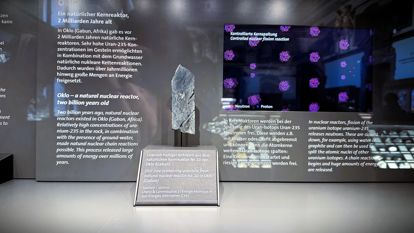 Ekspozycja muzealna z fragmentem z naturalnego reaktora jądrowego z Oklo, położonym na podstawce. Próbka jest otoczona szczegółowymi opisami naukowymi w kilku językach oraz ekranem prezentującym wizualizację rozmieszczenia neutronów i plutonu w reaktorze.