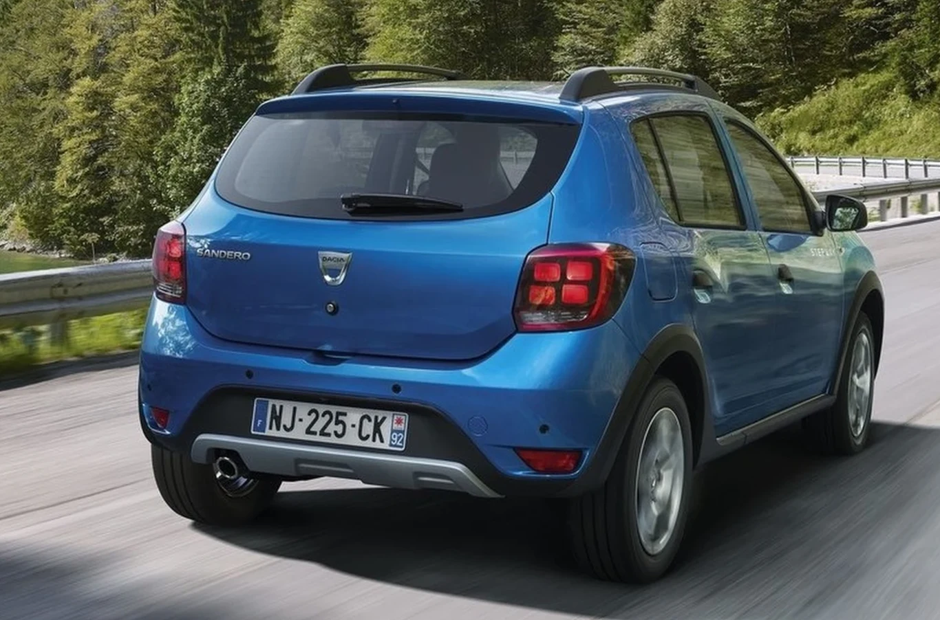 Niebieski samochód typu crossover Dacia Sandero jedzie asfaltową drogą otoczoną zielonym lasem, widoczny z tyłu z francuską tablicą rejestracyjną.
