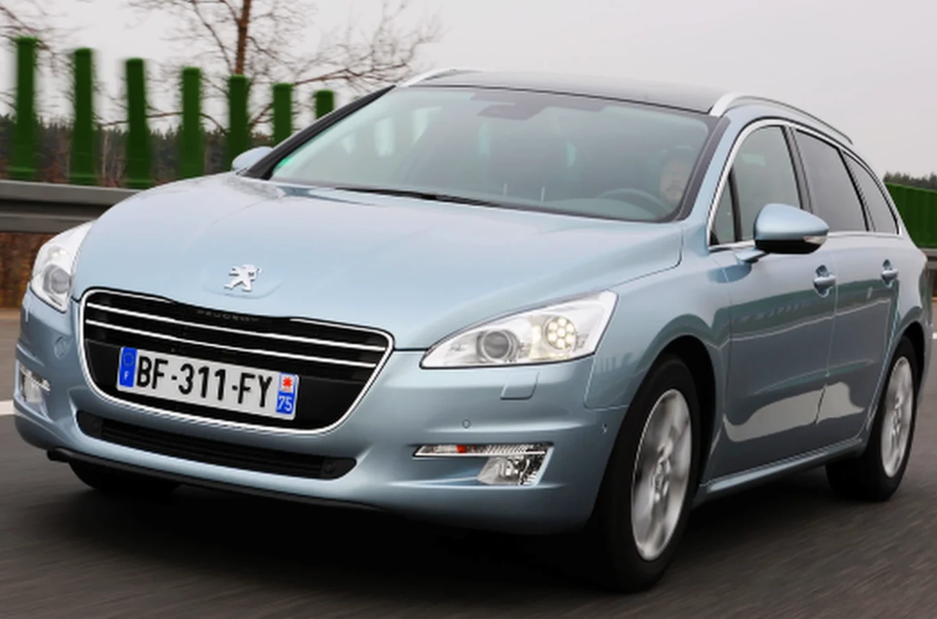 Jasnoniebieski samochód osobowy kombi Peugeot jedzie po asfaltowej drodze, na pierwszym planie wyraźnie widoczne reflektory, atrapę chłodnicy z logo marki oraz francuską tablicę rejestracyjną.