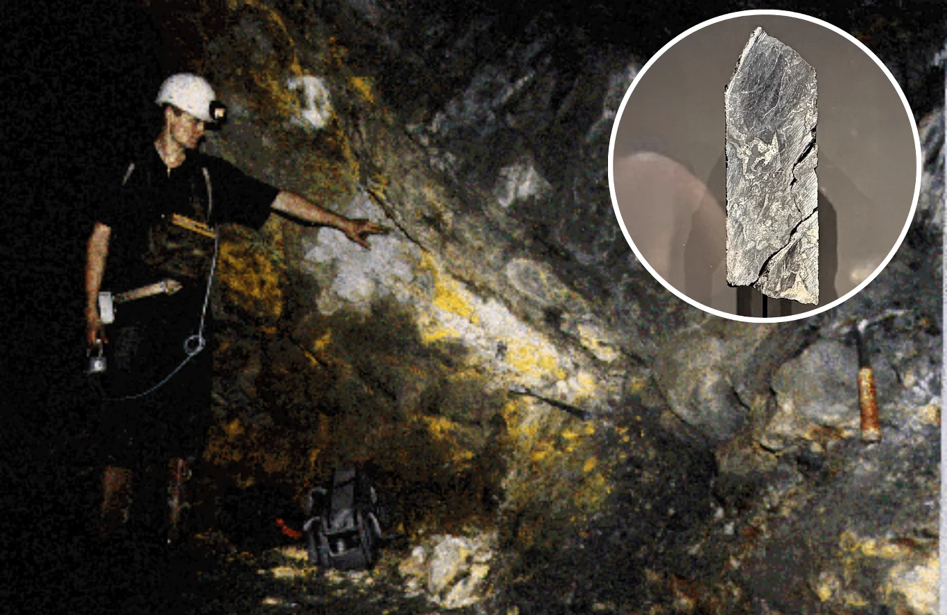 Górnik w kasku z latarką wskazuje fragment ściany kopalni z wyraźnymi żyłami minerałów, w prawym dolnym rogu znajduje się młotek geologiczny, a wstawka pokazuje powiększenie fragmentu skały wydobytego z tej warstwy.