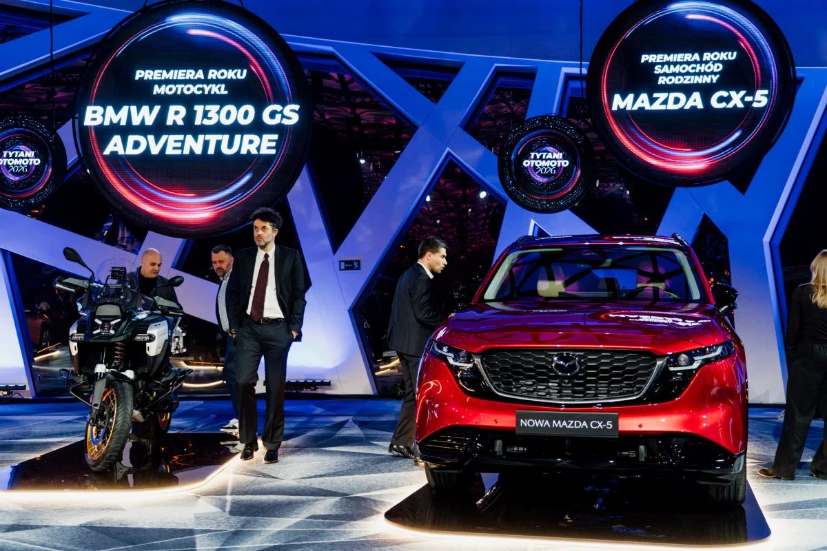 Motocykl BMW R 1300 GS Adventure i czerwony samochód Mazda CX-5 ustawione na scenie podczas prezentacji motoryzacyjnej, w tle świetlne panele LED z informacjami o premierach, kilka osób rozmawiających obok pojazdów.