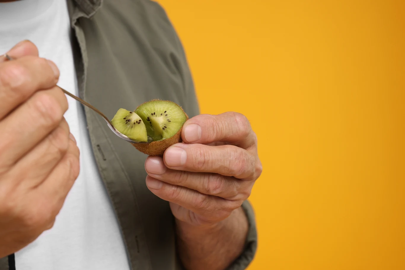 Osoba trzymająca w dłoni połówkę kiwi i nabierająca miąższ owocu łyżeczką na tle żółtej ściany.