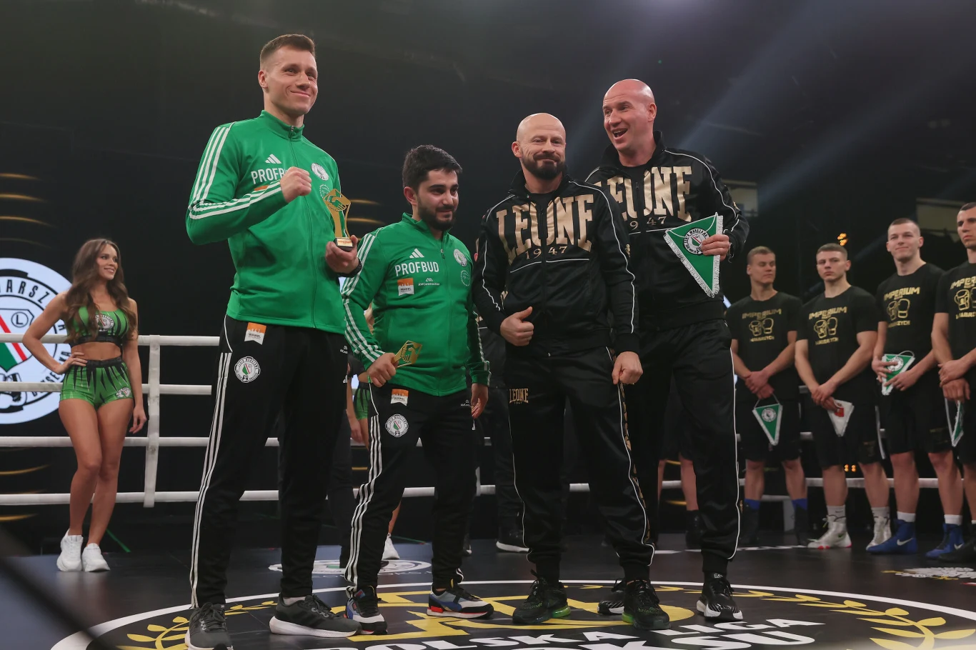 Czterech mężczyzn ubranych w sportowe stroje stoi na środku ringu, dwóch w zielonych dresach, dwóch w czarnych, trzymając puchary i statuetki, w tle grupy zawodników oraz hostessy w sportowych strojach.