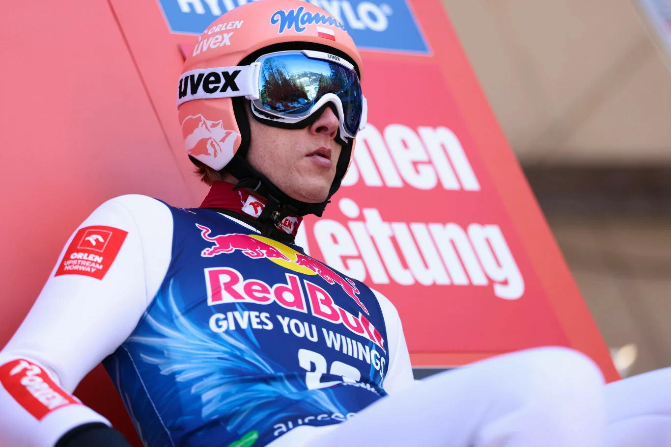 Skoczek narciarski w niebieskim kombinezonie z logo Red Bull, w kasku i goglach, siedzący na belce startowej podczas zawodów, w tle czerwony baner z białym napisem.