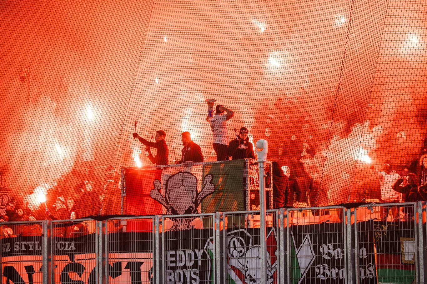 Tłum kibiców piłkarskich trzymających race świetlne, otoczony gęstym, pomarańczowym dymem i flagami z klubowymi emblematami oraz czaszką w centralnym punkcie trybuny.