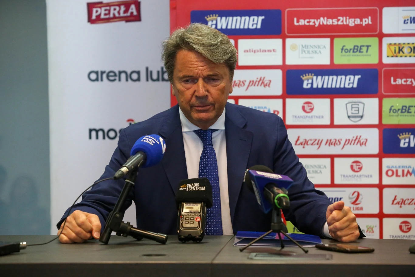 Mężczyzna w garniturze siedzi przy stole podczas konferencji prasowej, przed nim znajdują się mikrofony kilku stacji radiowych, w tle widoczna jest ściana z logotypami sponsorów i partnerów sportowych.