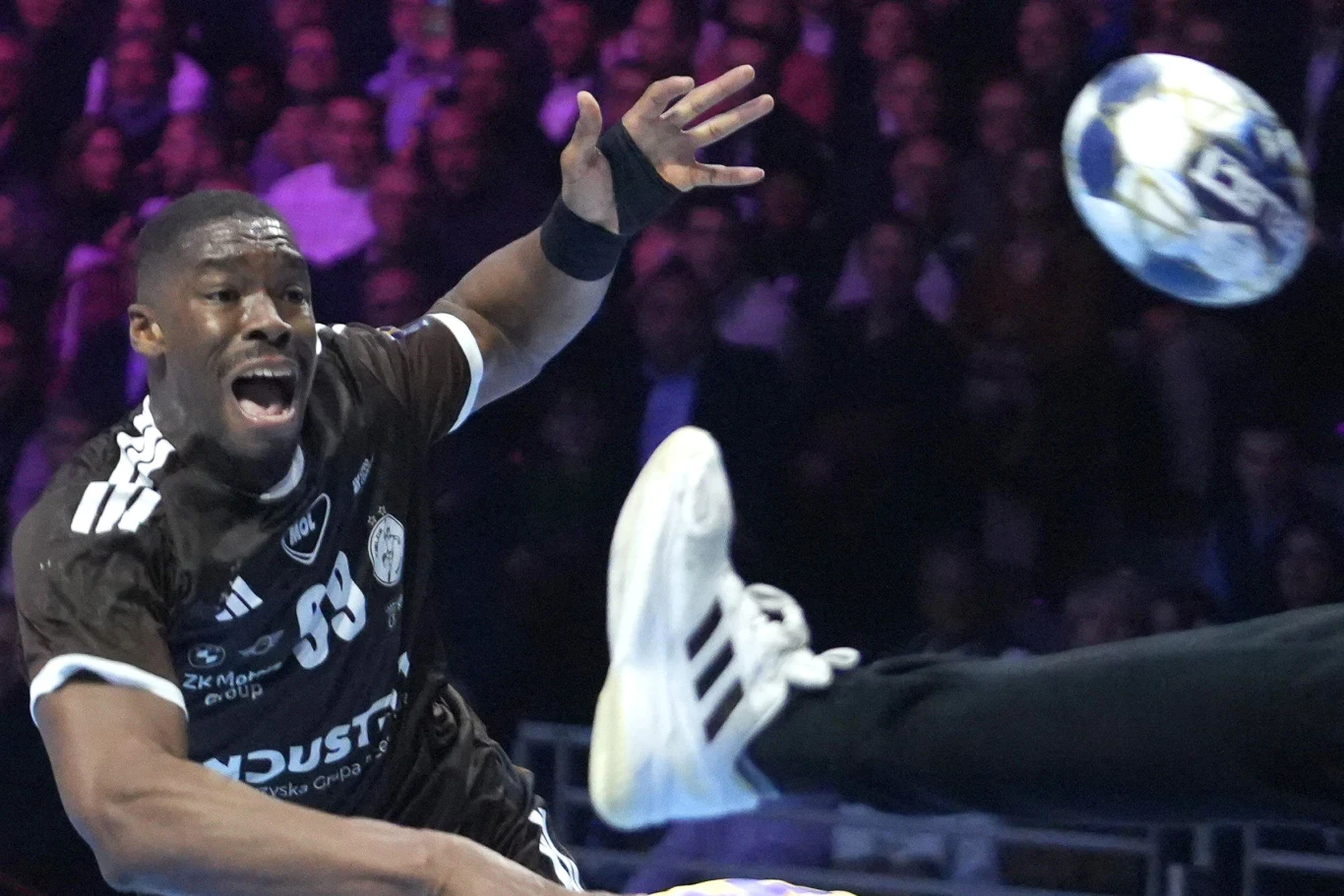 Zawodnik w czarnej koszulce z numerem 39 dynamicznie rzuca piłką ręczną w stronę bramki, jego twarz wyraża skupienie i wysiłek, tuż obok widoczna jest noga bramkarza próbującego zablokować strzał.