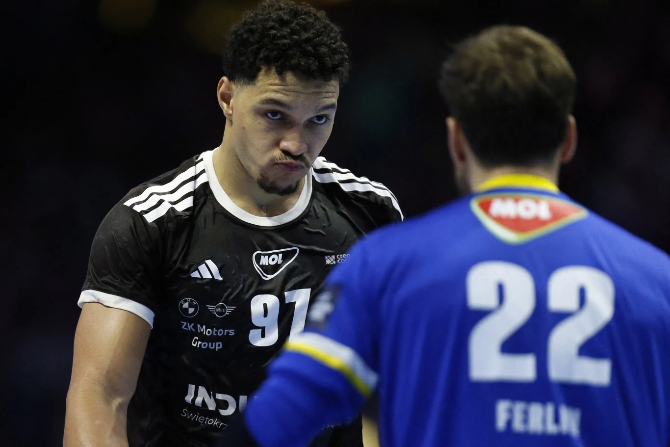 Dwóch szczypiornistów podczas meczu, zawodnik w czarnej koszulce z numerem 97 patrzy poważnie na przeciwnika ubranego w niebiesko-żółtą koszulkę z numerem 22, widoczne intensywne emocje i rywalizacja sportowa.