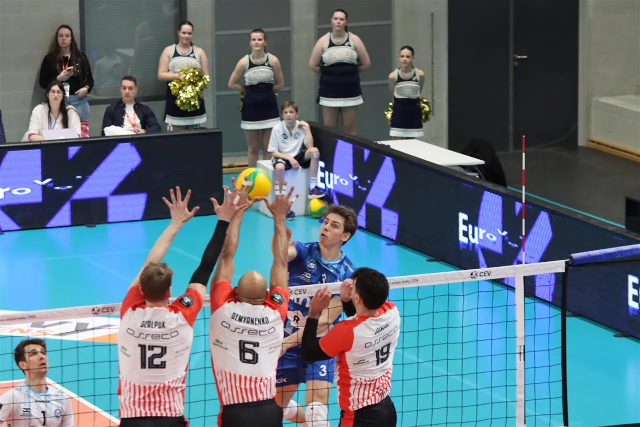 Zawodnik w niebieskim stroju wykonuje atak siatkarski nad siatką, podczas gdy trzech rywali w biało-czerwonych koszulkach próbuje zablokować piłkę, w tle cheerleaderki stojące przy bandach reklamowych.