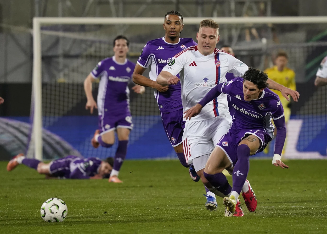 Dwóch piłkarzy walczących o piłkę podczas dynamicznego meczu, zawodnik w białym stroju próbuje zatrzymać przeciwnika w fioletowym stroju, w tle widoczni pozostali zawodnicy oraz bramkarz przy bramce.