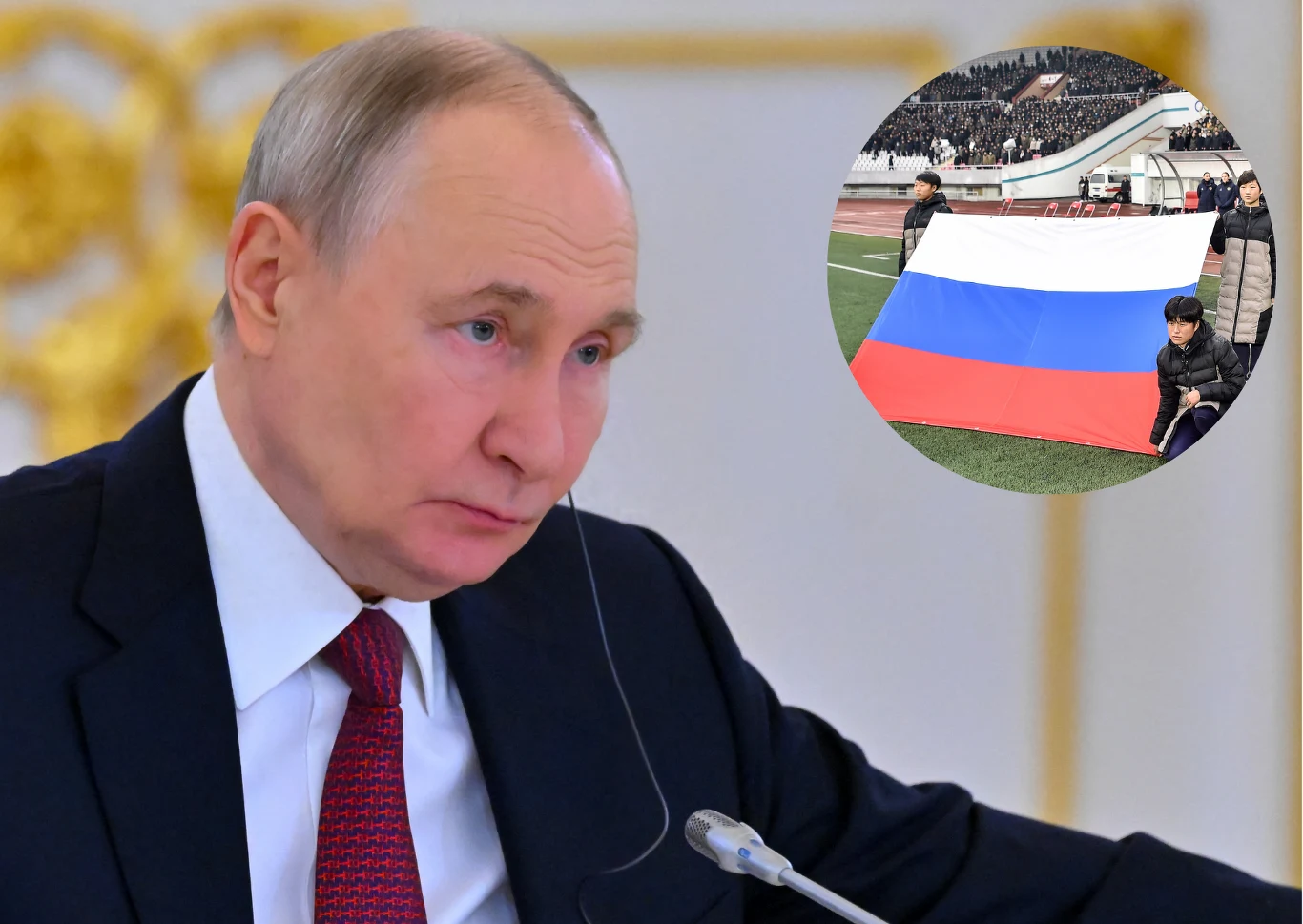 Mężczyzna w garniturze i czerwonym krawacie siedzi przy mikrofonie z poważnym wyrazem twarzy, w rogu widoczna jest duża rosyjska flaga trzymana przez kilka osób na stadionie.