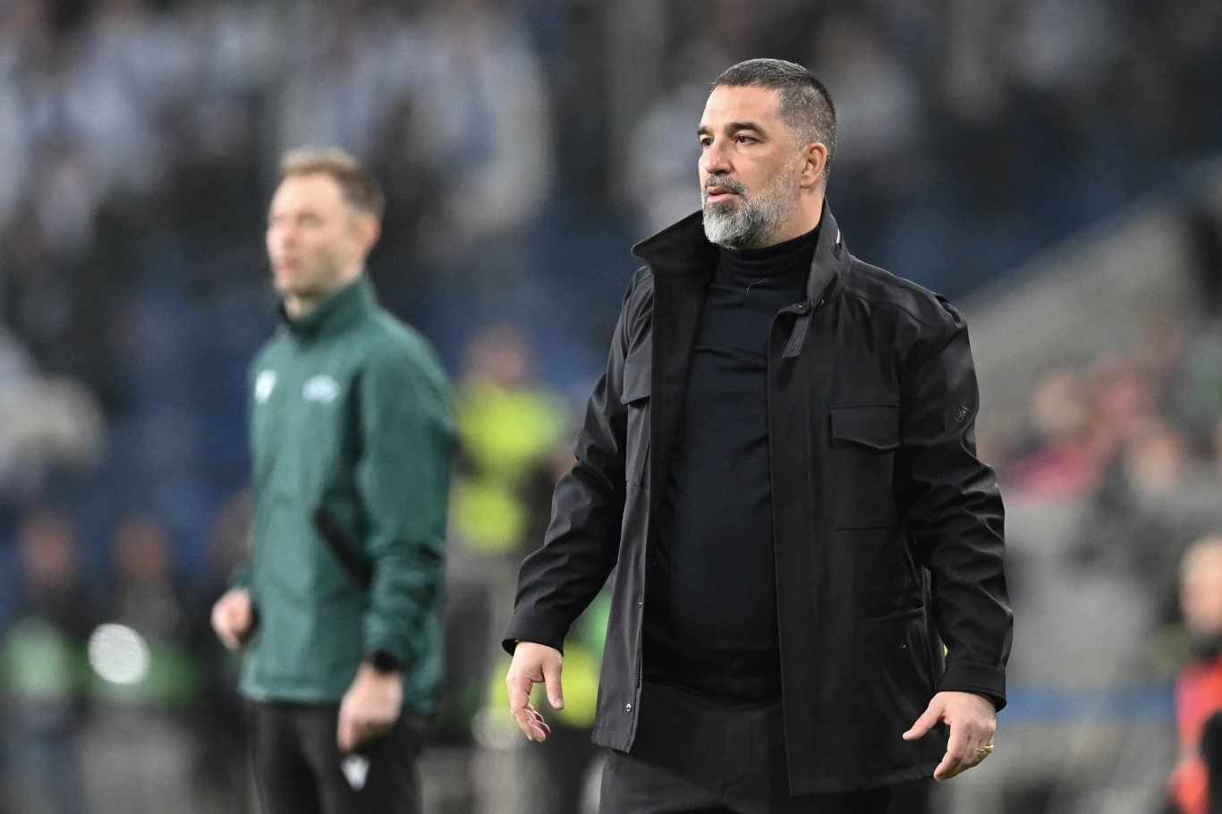 Mężczyzna w czarnym ubraniu stoi na pierwszym planie na boisku sportowym, z wyrazem skupienia na twarzy; w tle inny mężczyzna w zielonej kurtce obserwuje wydarzenia na boisku.