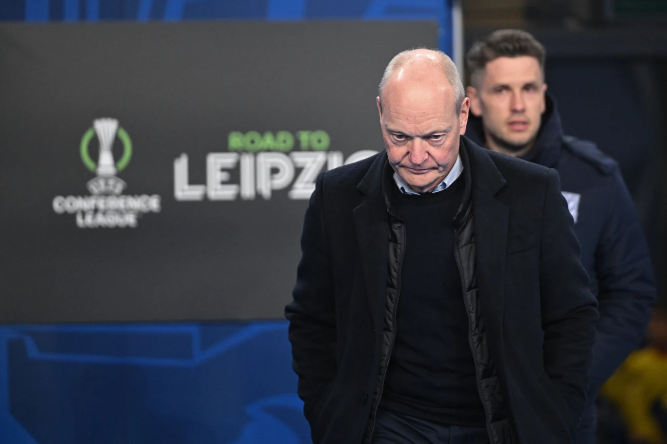 Starszy mężczyzna w ciemnej marynarce i swetrze z poważnym wyrazem twarzy stoi na tle tablicy z napisem „ROAD TO LEIPZIG” i logo Ligi Konferencji Europy UEFA, za nim młodszy mężczyzna w kurtce sportowej.