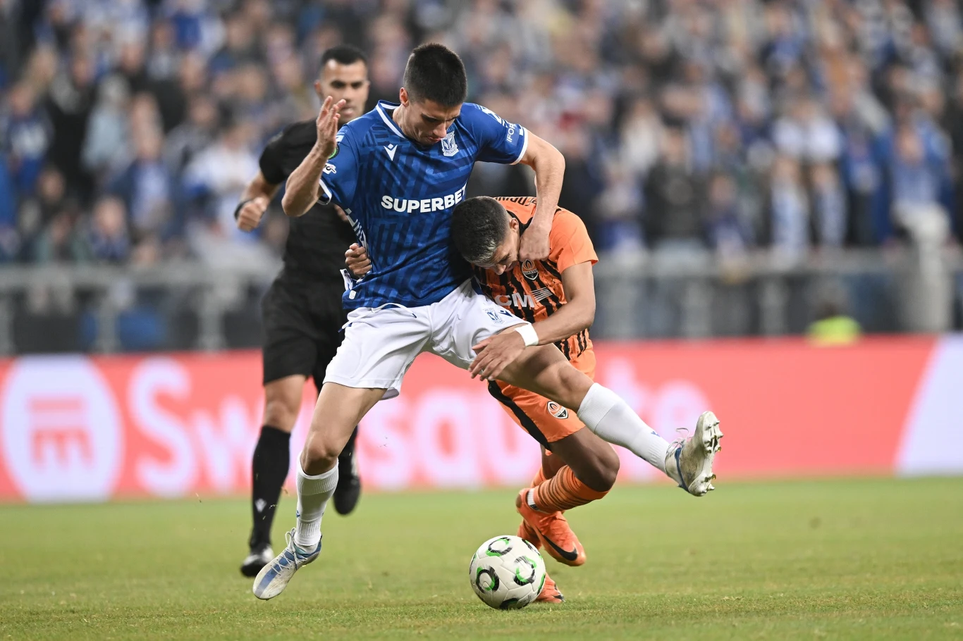 Dwóch piłkarzy w dynamicznej walce o piłkę, jeden ubrany w niebieską koszulkę i białe spodenki, drugi w pomarańczowym stroju; w tle sędzia oraz rozmyta publiczność na trybunach.