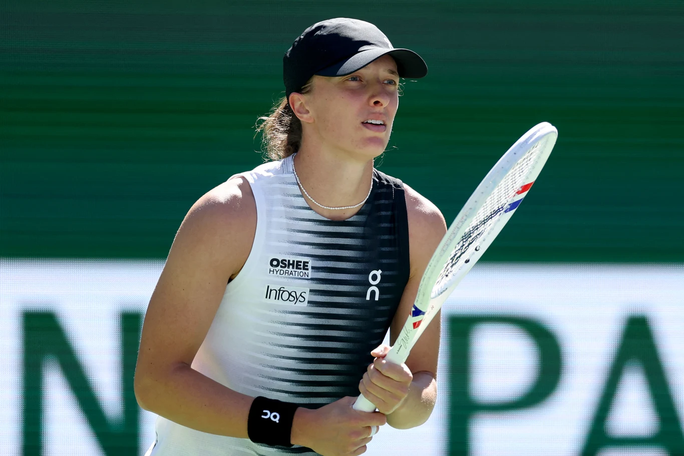 Tenisistka w sportowym stroju i czapce z daszkiem, trzymająca rakietę tenisową i skoncentrowana podczas meczu na tle kortu tenisowego.