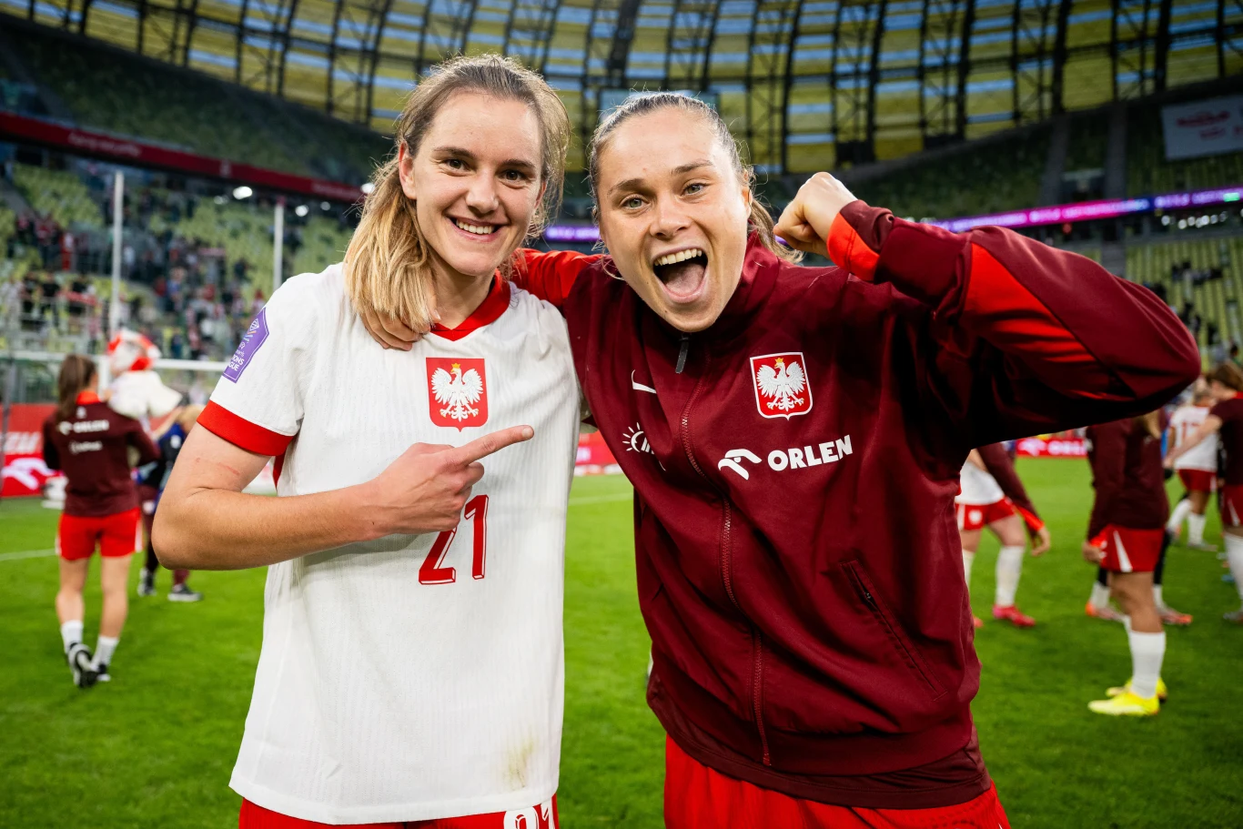Paulina Tomasiak oraz Ewa Pajor Dwie piłkarki reprezentacji Polski uśmiechają się i pozują razem na boisku po meczu, jedna z nich wskazuje na drugą, obie ubrane są w stroje narodowe, a w tle widać stadion i innych członków drużyny.