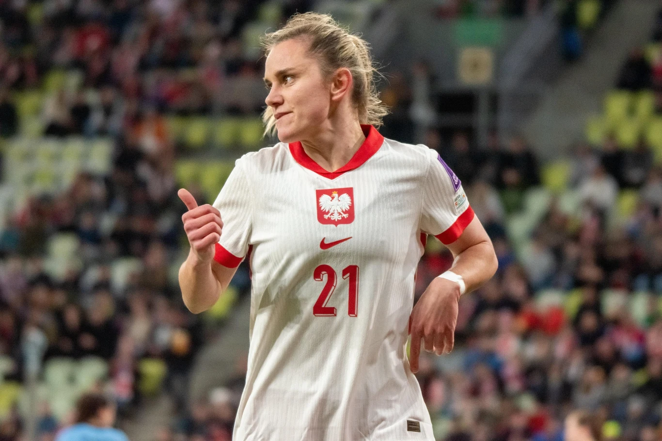 Piłkarka w białej koszulce z numerem 21 oraz orłem na piersi wykonuje gest uniesionego kciuka, wokół widać trybuny wypełnione kibicami.