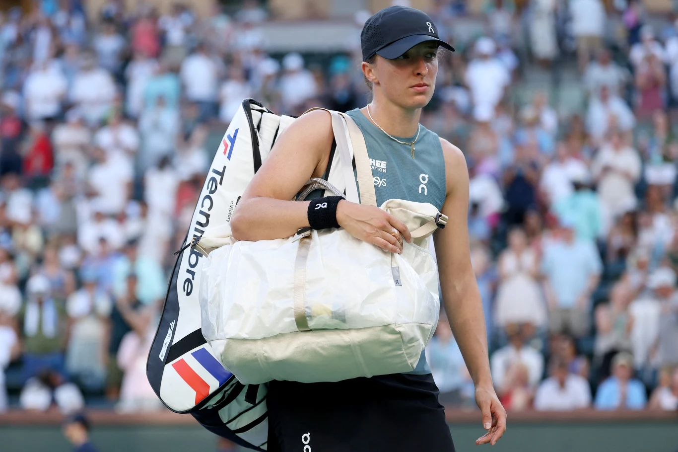 Tenisistka w sportowym stroju i czapce z daszkiem niesie dwie torby sportowe na tle rozmytego tłumu widzów na trybunach kortu tenisowego.