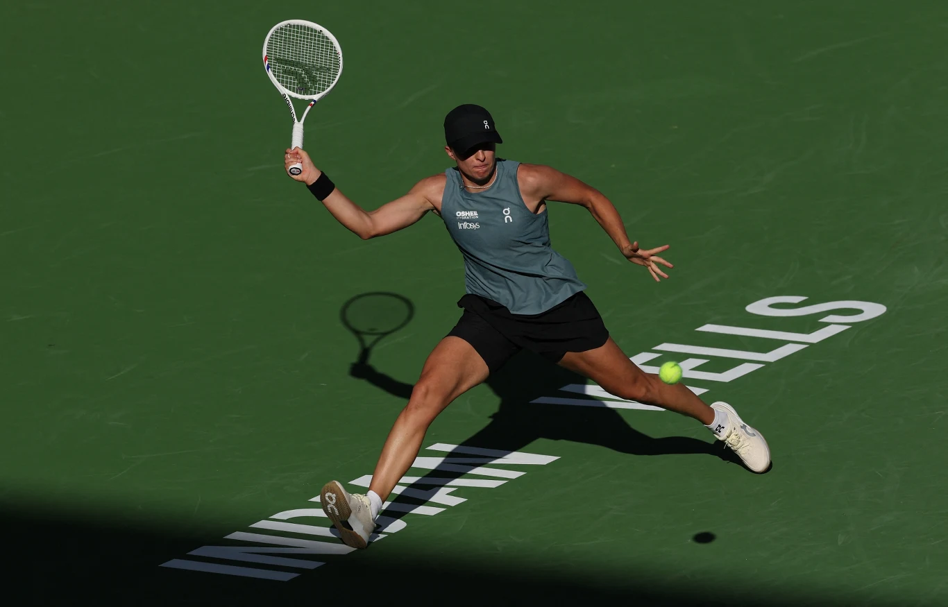 Tenisistka w sportowym stroju podczas dynamicznego ruchu wyciąga rakietę do odbicia piłki na zielonym korcie oznaczonym napisem INDIAN WELLS, cień sportowca i rakiety widoczny na korcie.