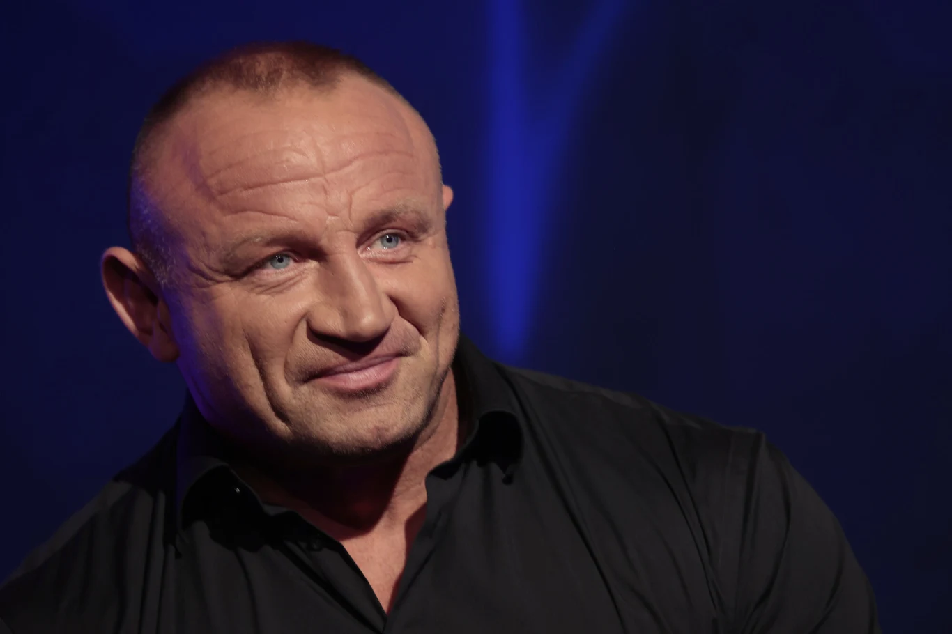 Uśmiechnięty, muskularny mężczyzna w czarnej koszuli na tle ciemnego, jednolitego tła.