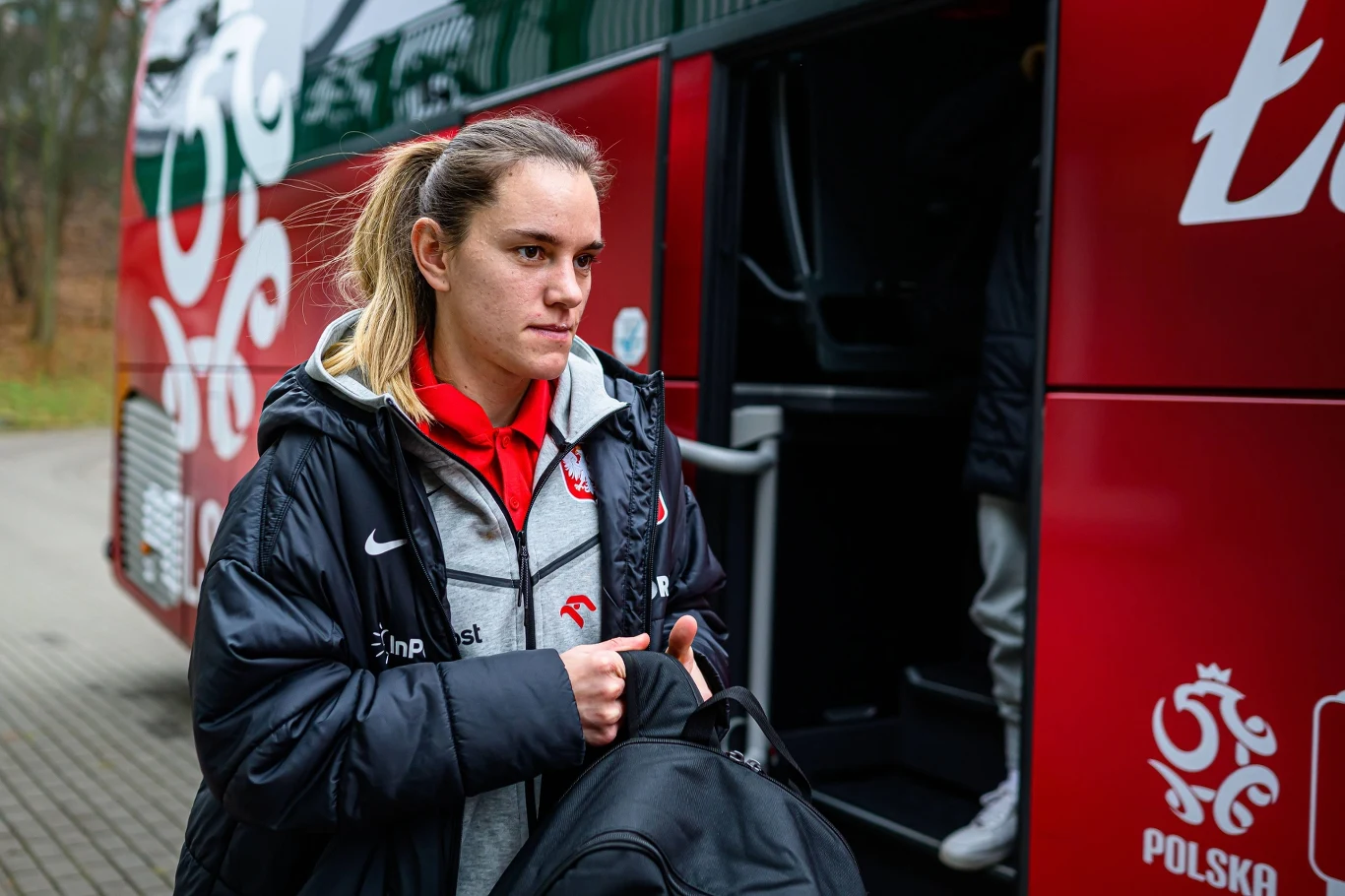 Młoda kobieta w sportowej kurtce z orzełkiem na piersi wychodzi z autokaru reprezentacji Polski, trzymając czarną torbę podróżną, w tle fragment pojazdu z napisem „Polska” i godłem narodowym.