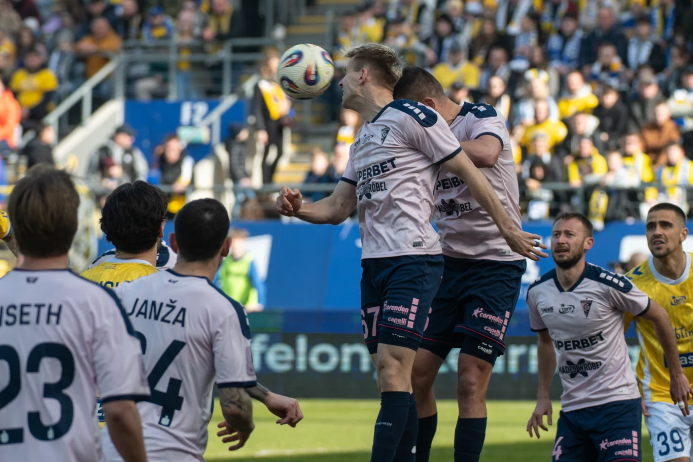 Grupa piłkarzy walczy o piłkę w powietrzu podczas meczu, kilku zawodników w białych koszulkach rywalizuje z piłkarzami w żółtych strojach, w tle widać trybuny pełne kibiców.