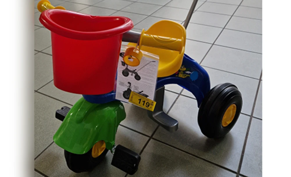 Kolorowy dziecięcy rowerek trójkołowy z czerwonym koszykiem z przodu, żółtym siedziskiem i kierownicą, stojący na płytkach podłogowych; do rowerka przymocowana jest etykieta z ceną 119 zł.