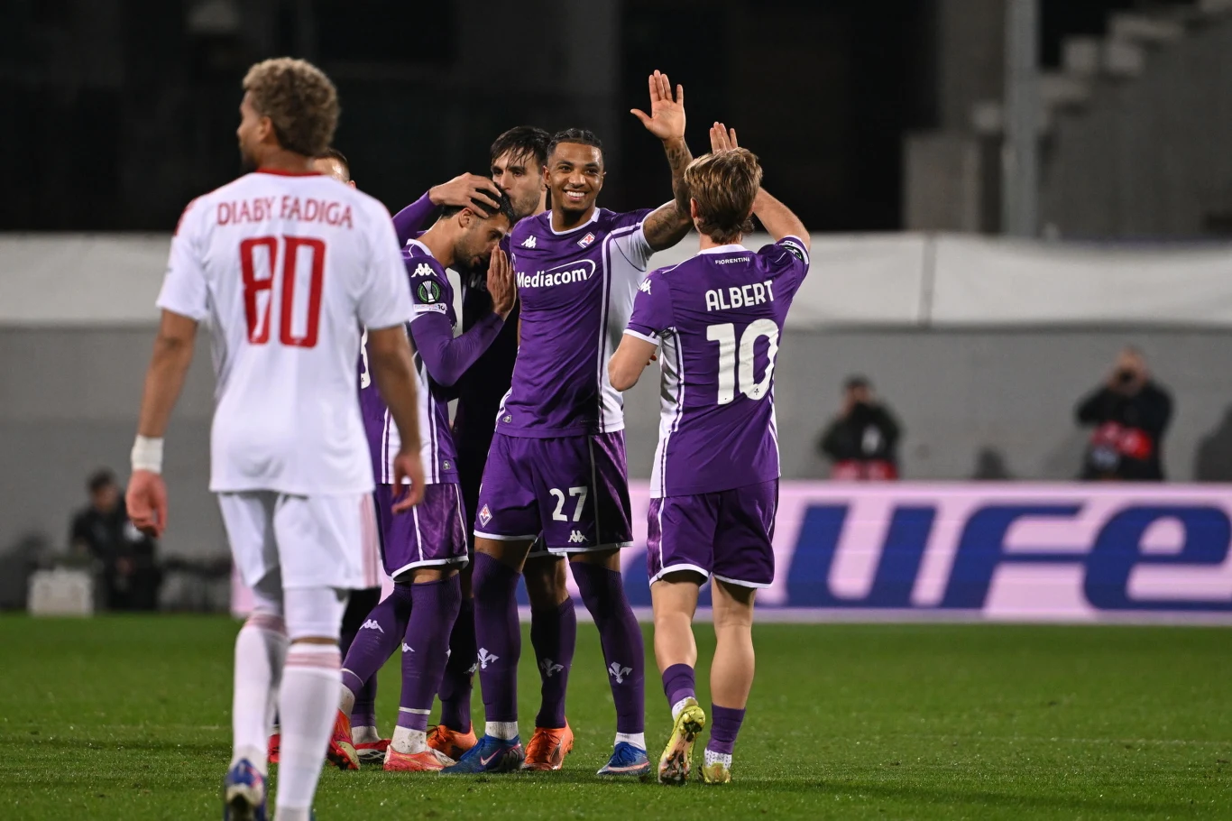 Grupa piłkarzy w fioletowych strojach świętuje zdobycie bramki na boisku piłkarskim, podczas gdy zawodnik drużyny przeciwnej w białym stroju z numerem 80 odchodzi w tle.