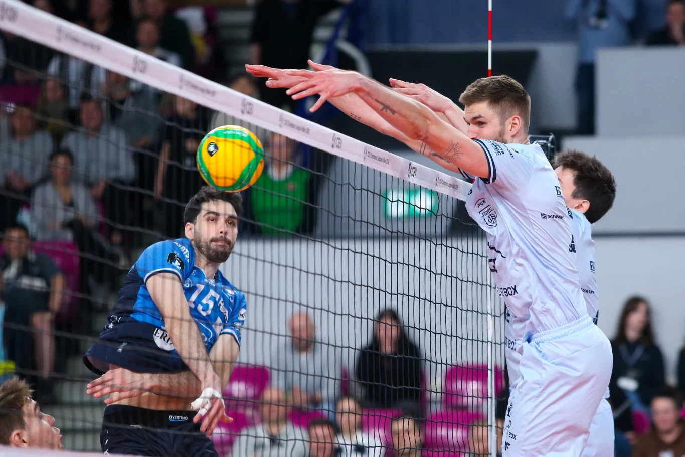 Dwóch siatkarzy w białych strojach wykonuje blok przy siatce, próbując zatrzymać atak zawodnika drużyny przeciwnej w ciemnoniebieskim stroju, który uderza piłkę w kierunku bloku; w tle widoczna publiczność na trybunach.