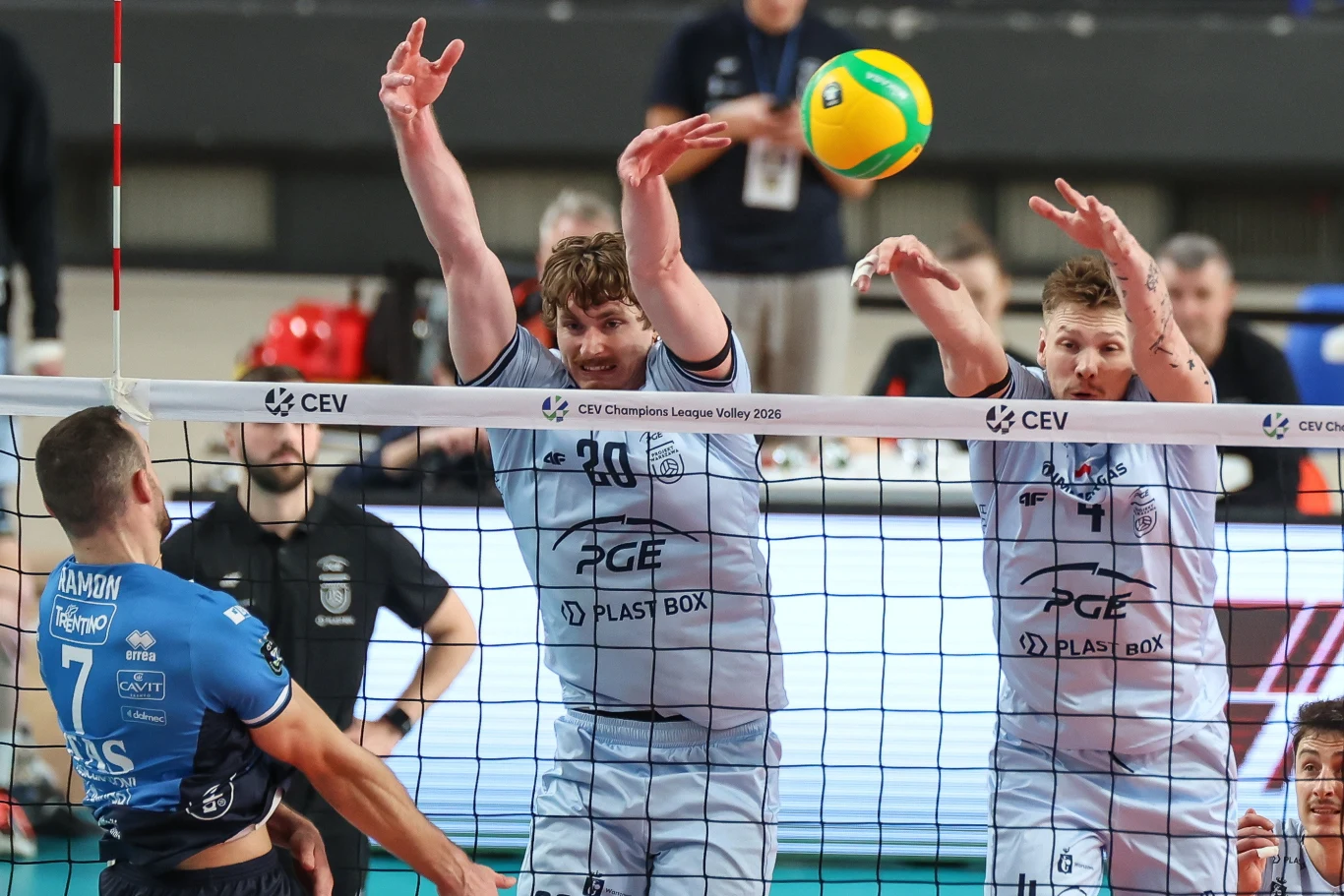 Dwóch siatkarzy drużyny w jasnoszarych strojach próbuje zablokować piłkę nad siatką, podczas gdy zawodnik przeciwnej drużyny w granatowym trykocie przygotowuje się do ataku.