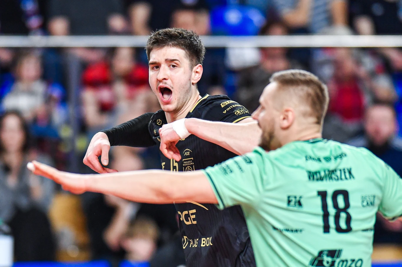 Dwóch siatkarzy podczas dynamicznej akcji meczowej, zawodnik w czarnym stroju wyraża silne emocje, drugi w zielonej koszulce z numerem 18 zwrócony bokiem; w tle rozmyta publiczność na trybunach.