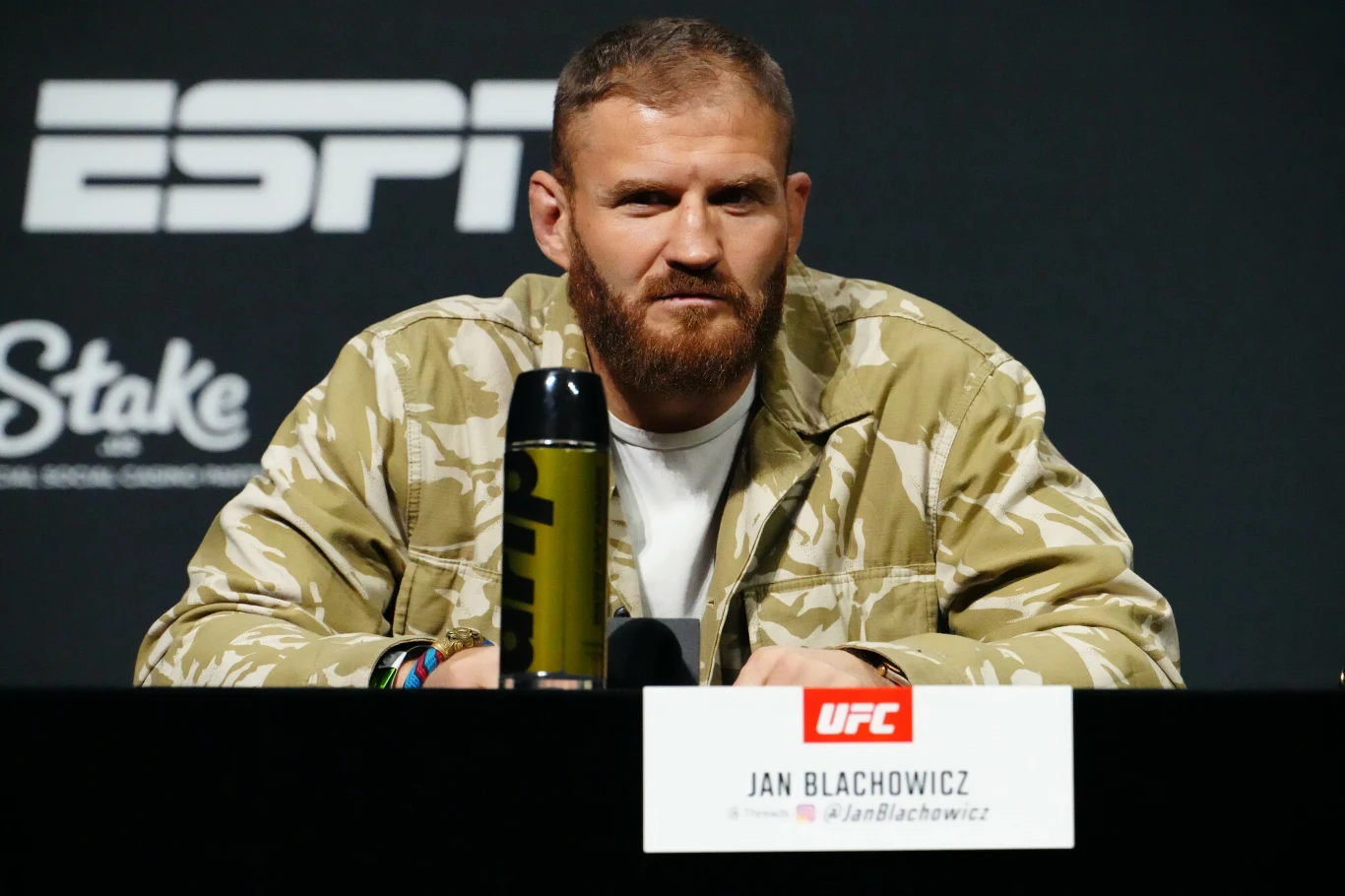 Mężczyzna z brodą w jasnej kurtce moro siedzi przy stole konferencyjnym na tle logotypów ESPN i UFC, przed nim stoi mikrofon oraz tabliczka z jego imieniem i nazwiskiem.