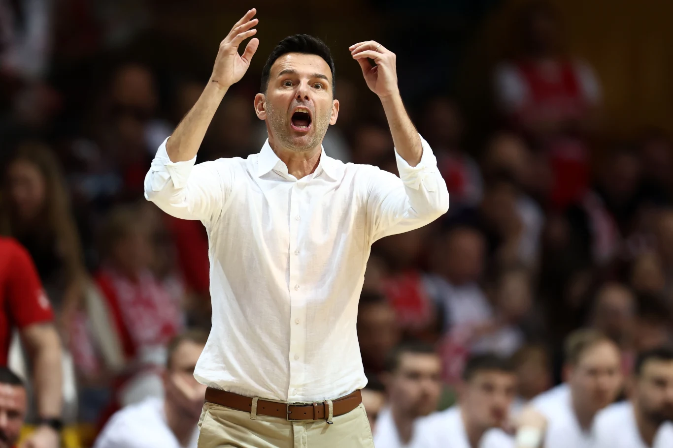 Mężczyzna w białej koszuli gestykuluje energicznie, stojąc naprzeciw dużej grupy ludzi siedzących na trybunach, wyrażając silne emocje.