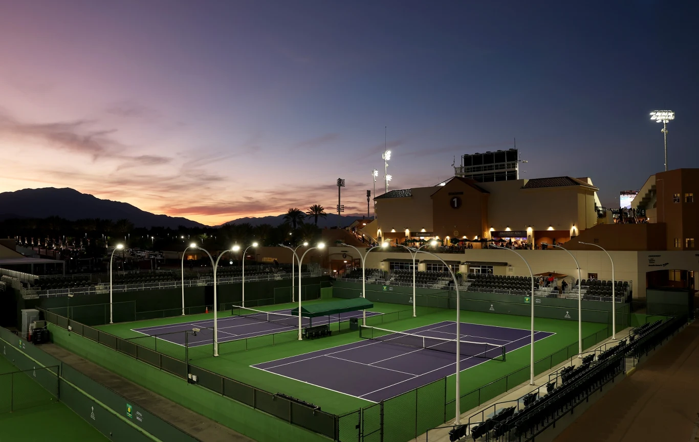 Oświetlone korty tenisowe otoczone trybunami, wieczorne niebo w tle z widocznym zachodem słońca oraz sylwetką budynków kompleksu sportowego.