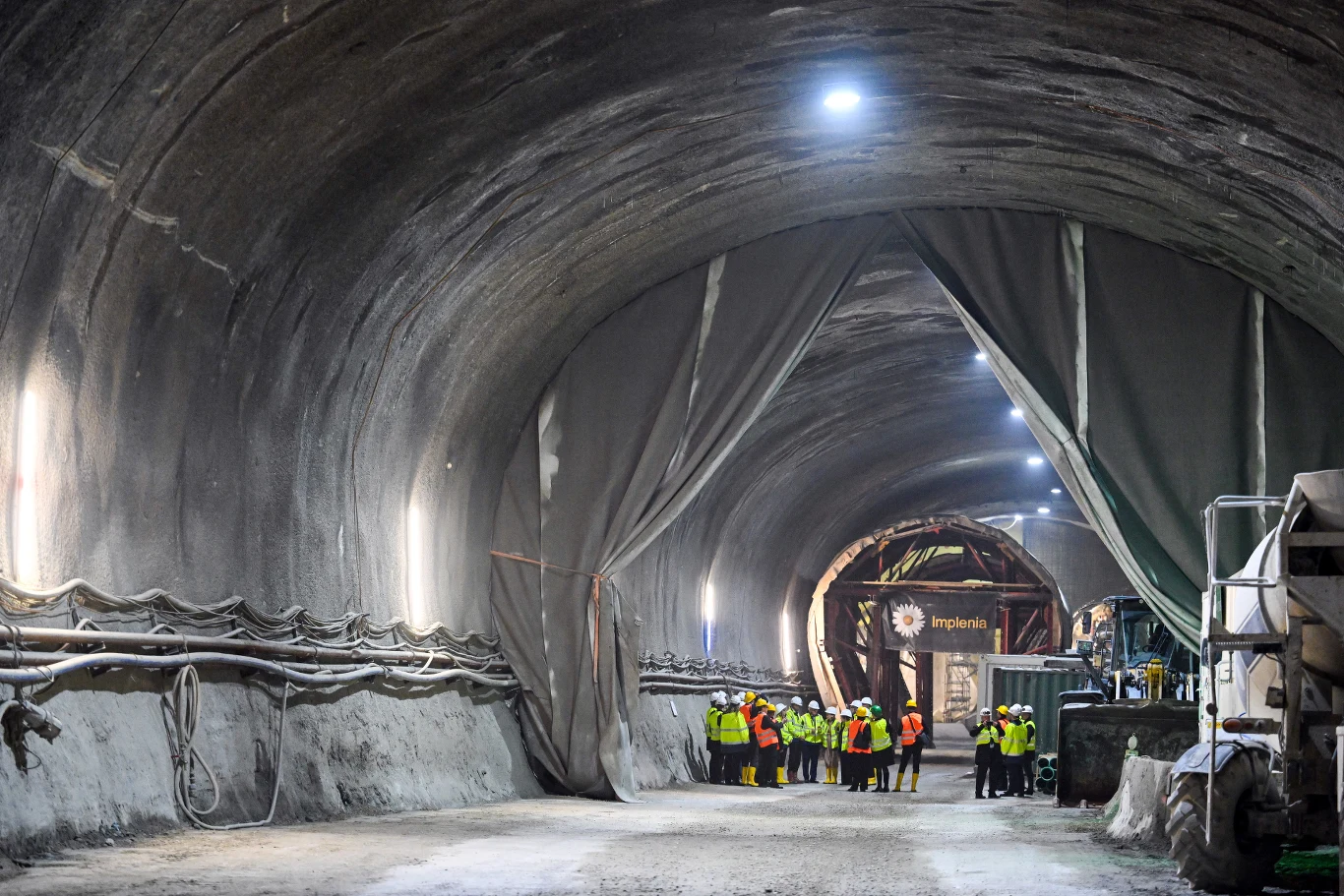 Brenner Base Tunnel. Grupa osób w żółtych kamizelkach ochronnych stoi wewnątrz dużego tunelu, oświetlonego sztucznym światłem, otoczona instalacjami oraz ciężkim sprzętem budowlanym.