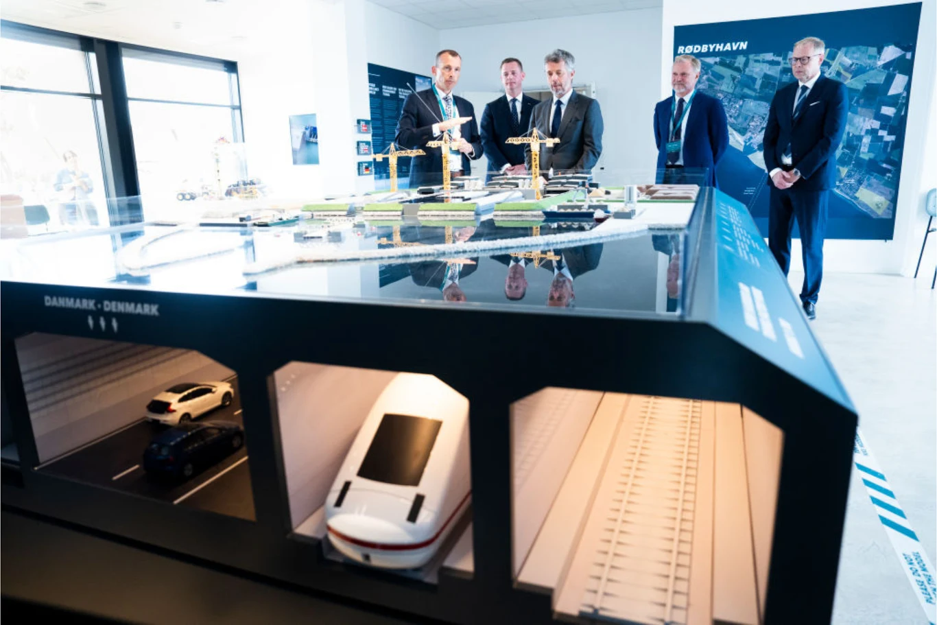 Tunelem pod Bałtykiem przejadą zarówno samochody, jak i pociągi. Model dużej infrastruktury tunelowej z widocznymi przekrojami drogowymi i kolejowymi, wokół którego stoi grupa elegancko ubranych mężczyzn w garniturach, omawiających projekt w nowoczesnym, jasnym wnętrzu wystawowym.