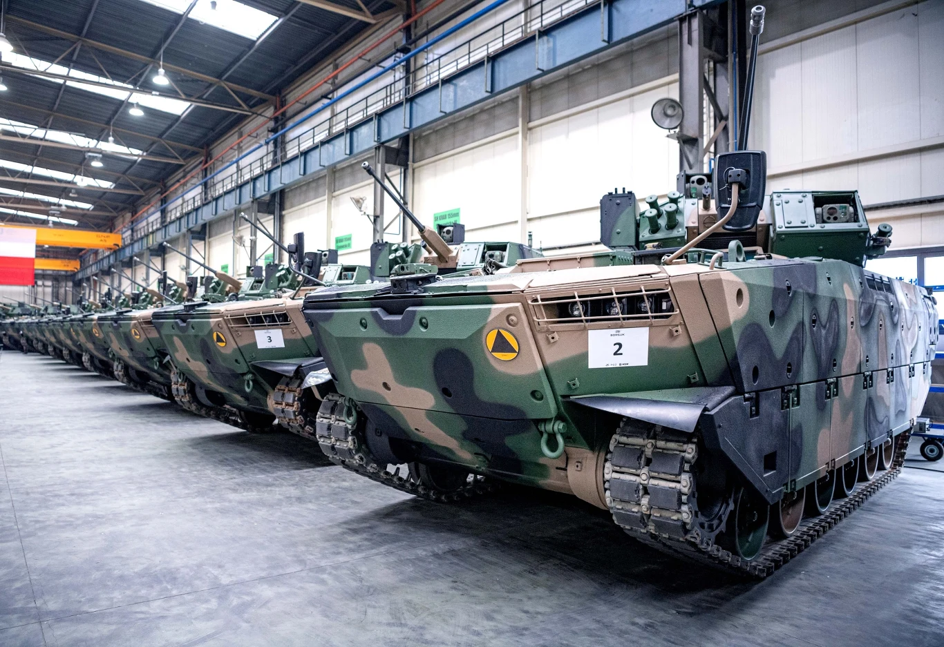 Rząd nowoczesnych bojowych wozów piechoty w kamuflażu, ustawionych w hangarze produkcyjnym, z widocznymi numerami identyfikacyjnymi na przednich pancerzach.