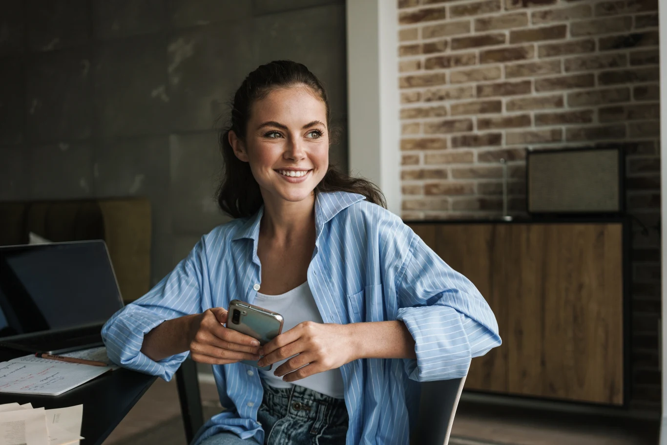 Błękitna koszula to idealny wybór na wiosnę Uśmiechnięta młoda kobieta w niebieskiej koszuli siedząca przy stole, trzymająca w dłoniach smartfon, z notatkami oraz laptopem obok siebie na stole; w tle drewniana komoda i ceglana ściana.