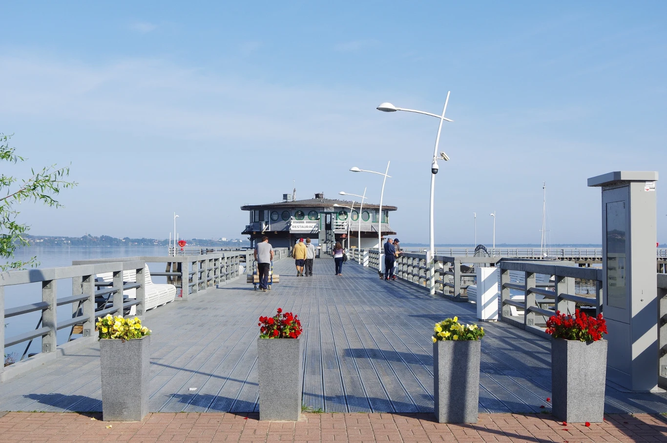 Spokojna alternatywa dla zatłoczonych kurortów nad Bałtykiem. Dlaczego warto odwiedzić Puck? Molo w Pucku prowadzące do budynku nad wodą, na którym spaceruje kilka osób, W tle widać spokojną tafle wody.