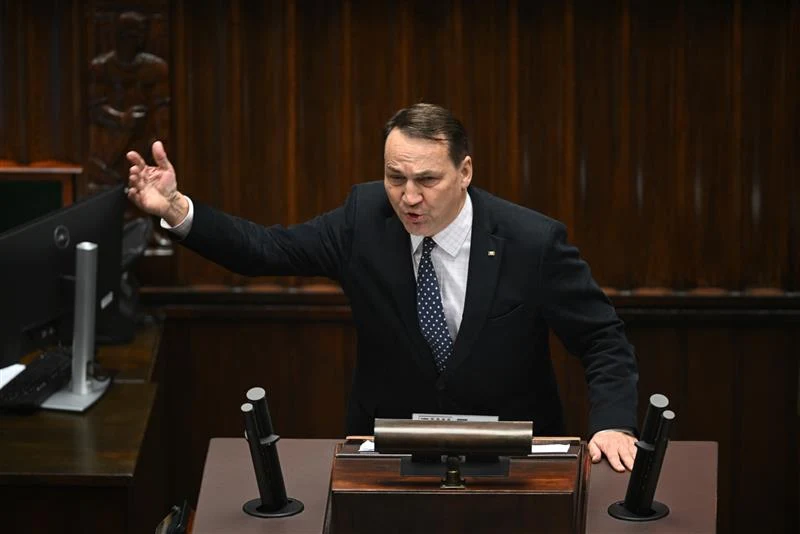 Mężczyzna w garniturze i krawacie gestykuluje ręką podczas przemówienia przy mównicy w sali obrad parlamentu.