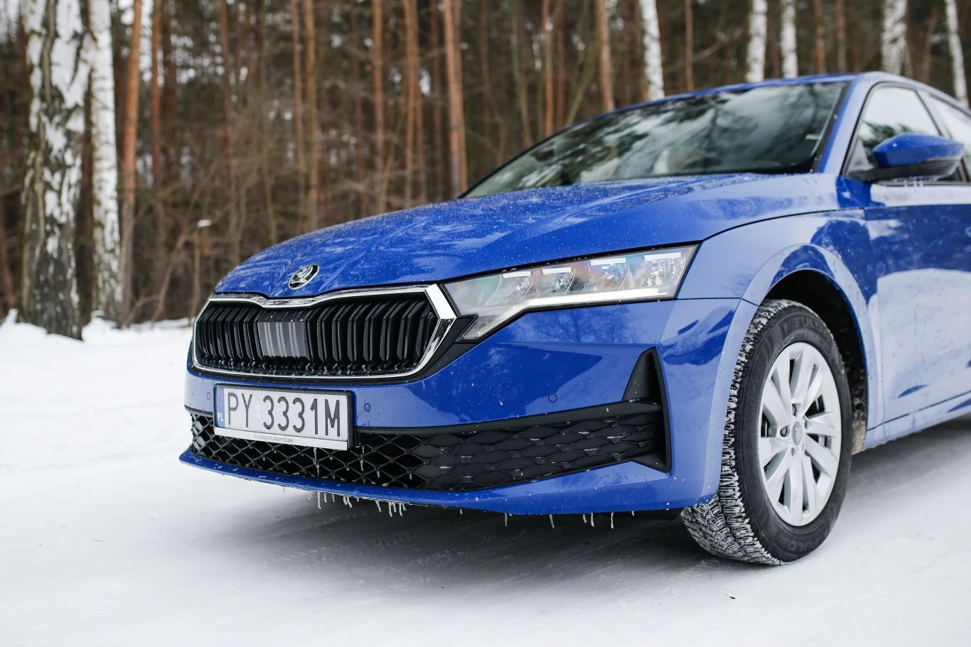 Niebieska Skoda zaparkowana na ośnieżonej drodze, z widocznymi soplami lodu na przednim zderzaku, w tle sosnowy las.
