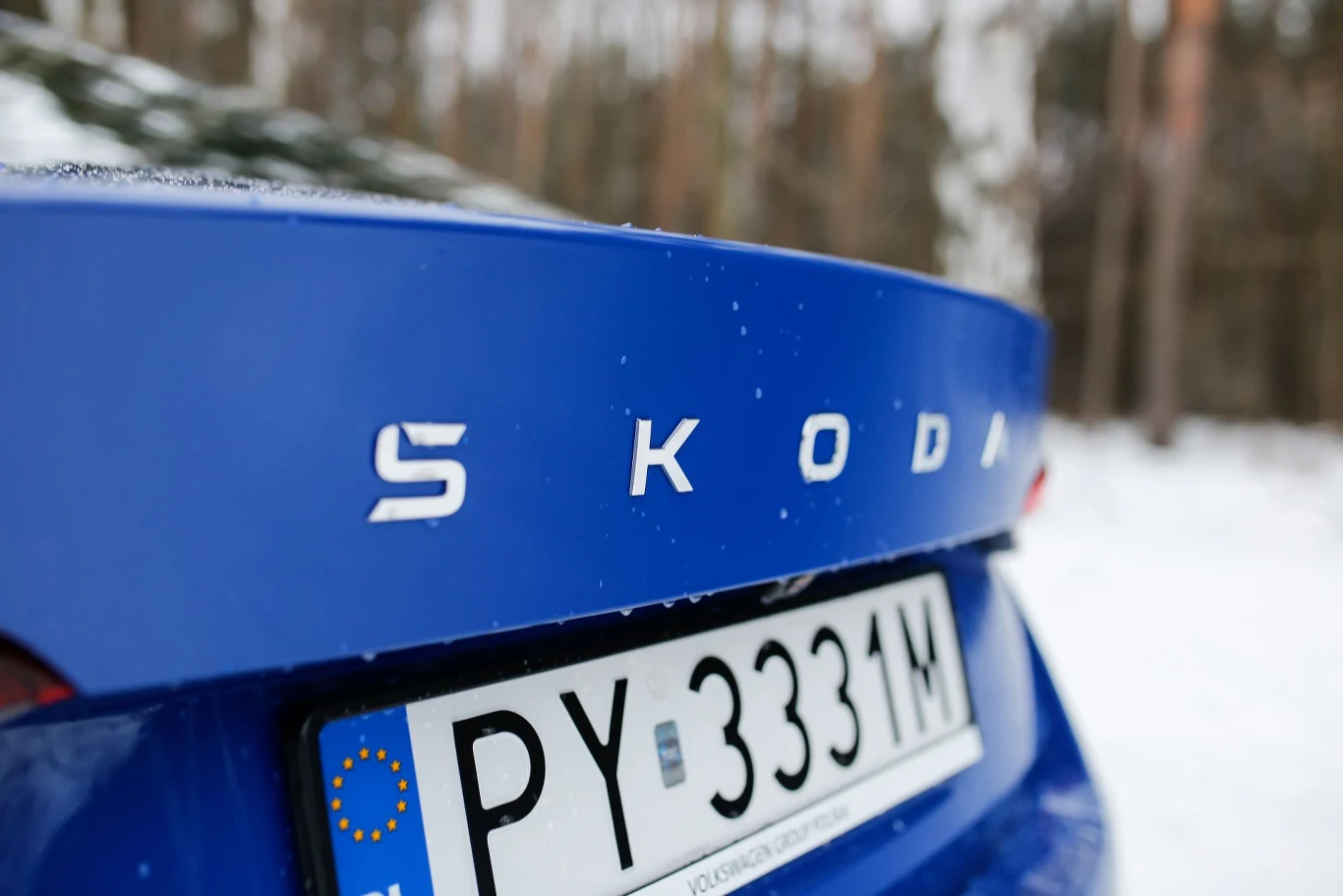Niebieski samochód marki Škoda z widocznym logo oraz polską tablicą rejestracyjną PY 3331M, w tle rozmyte drzewa i śnieg.