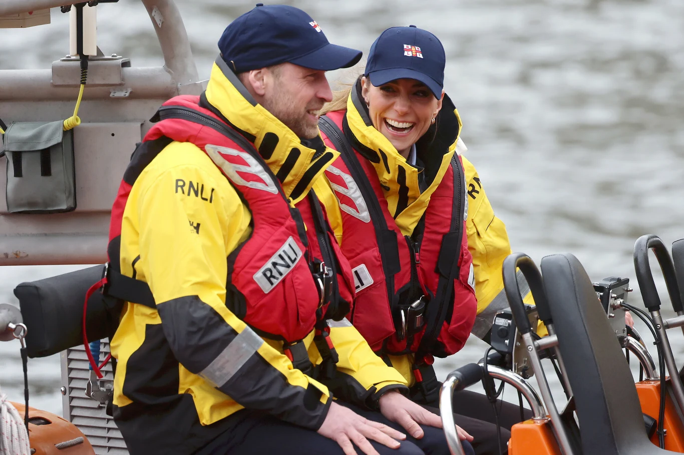 Dwoje ludzi ubranych w żółte kurtki i czerwone kamizelki ratunkowe RNLI siedzi na łodzi ratunkowej, uśmiechając się do siebie.