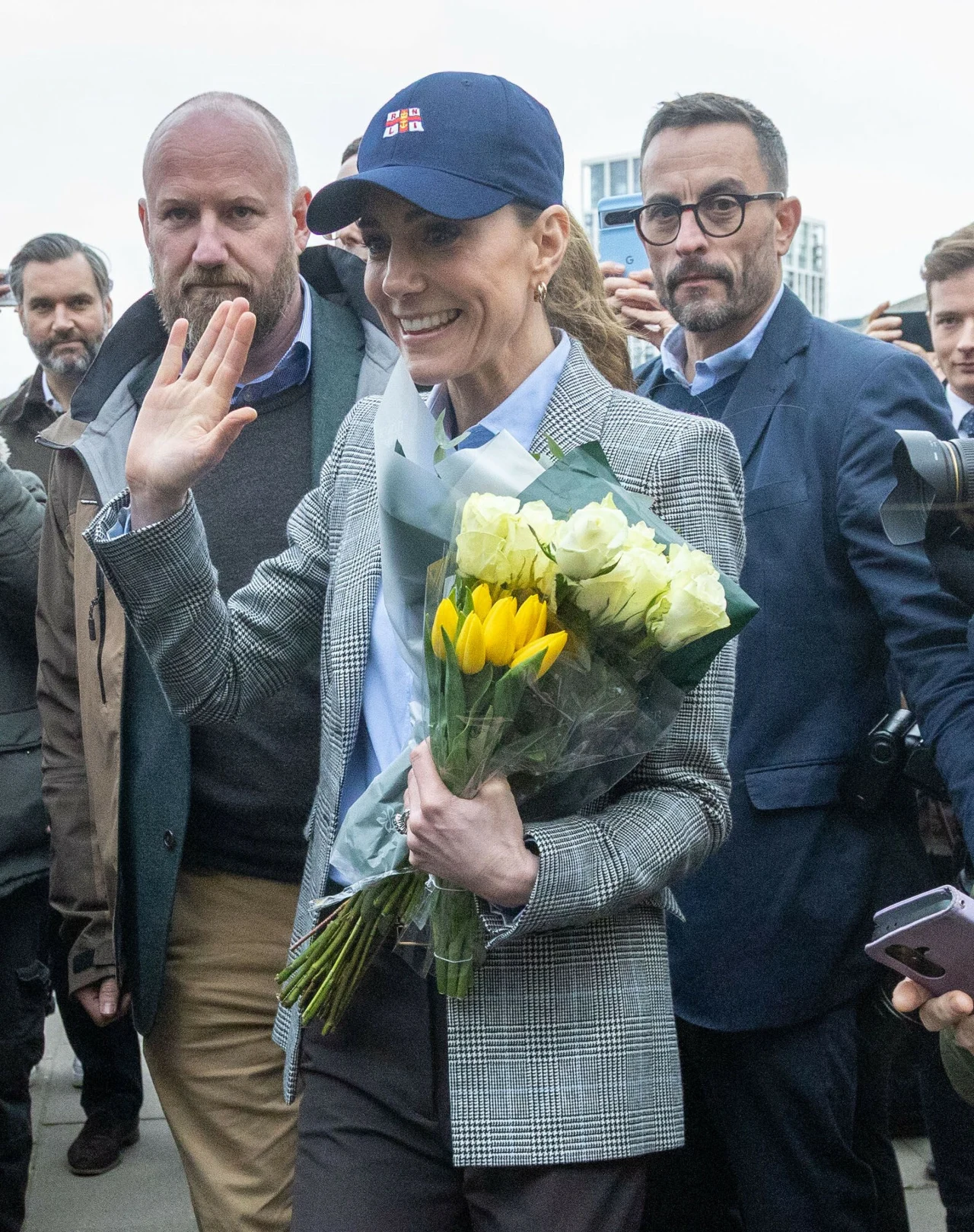 Uśmiechnięta kobieta w granatowej czapce z daszkiem trzyma bukiet żółtych i białych kwiatów, gestykulując ręką w kierunku zgromadzonych ludzi, otoczona przez kilku mężczyzn w garniturach.