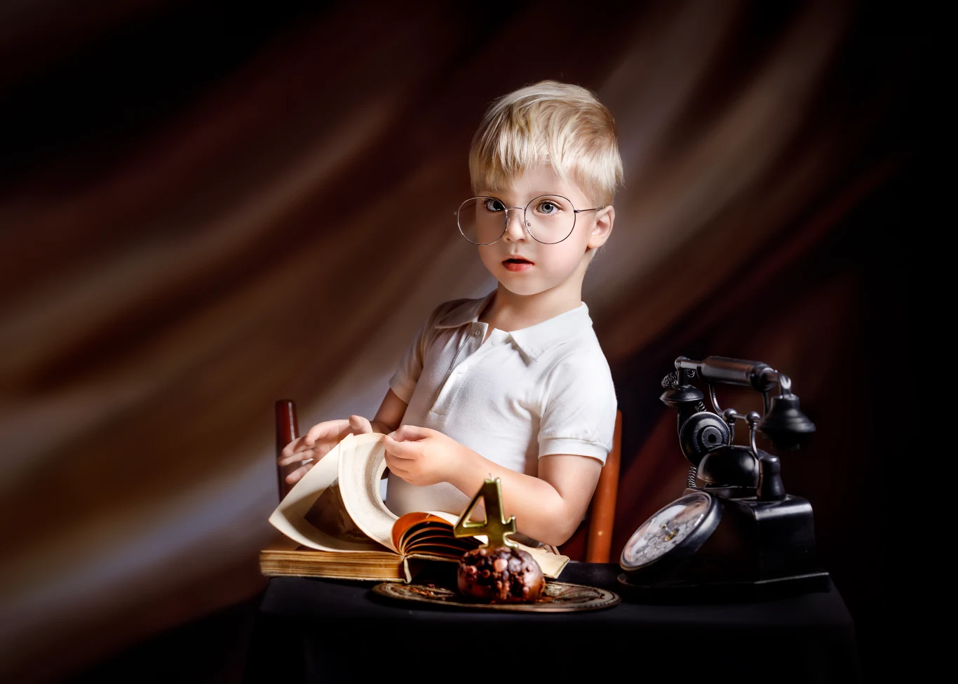 Chłopiec w okularach siedzi przy stole, przewracając strony książki; obok znajduje się zabytkowy telefon, zegar i talerz z babeczką oraz świeczką w kształcie cyfry 4.