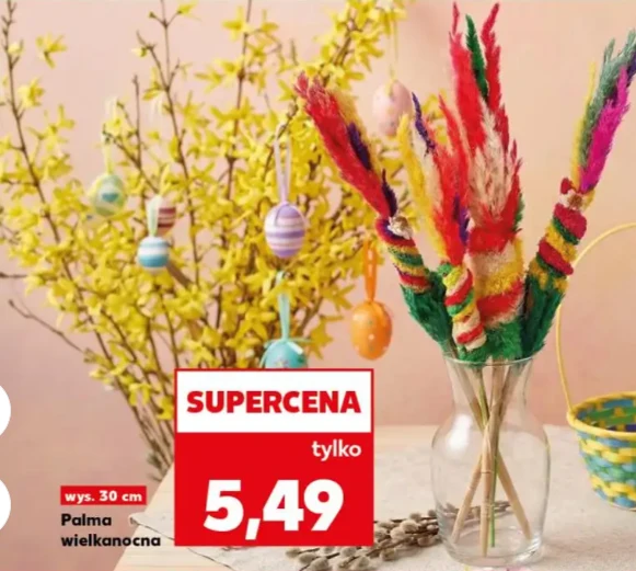 Wazon z kolorowymi palmami wielkanocnymi ustawiony na stole, w tle rozkwitająca gałązka forsycji ozdobiona pastelowymi, pisankowymi jajkami oraz żółty koszyk, na pierwszym planie widoczna cena promocyjna.
