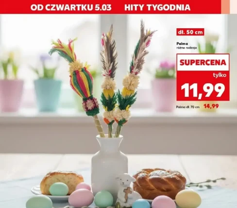Biała waza z kolorowymi palmami wielkanocnymi ustawiona centralnie na stole, wokół niej pastelowe jajka, baranek cukrowy oraz tradycyjne wypieki wielkanocne, w tle rozmyte doniczki z kwiatami na parapecie.